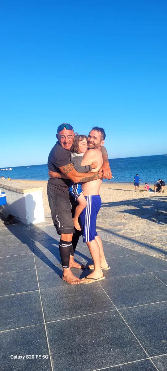 Two men and a child hug and laugh by the sea