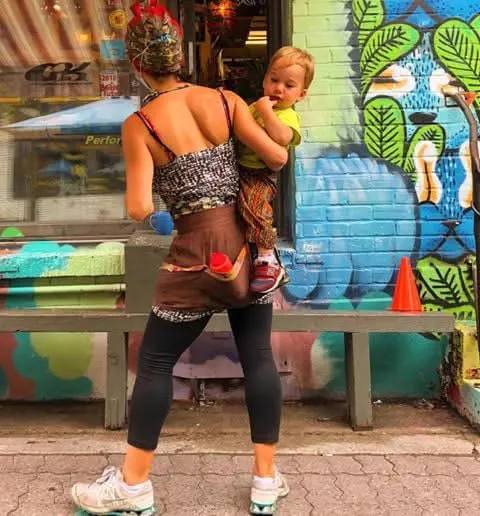 A woman seen from behind carries a baby in front of a colourful mural