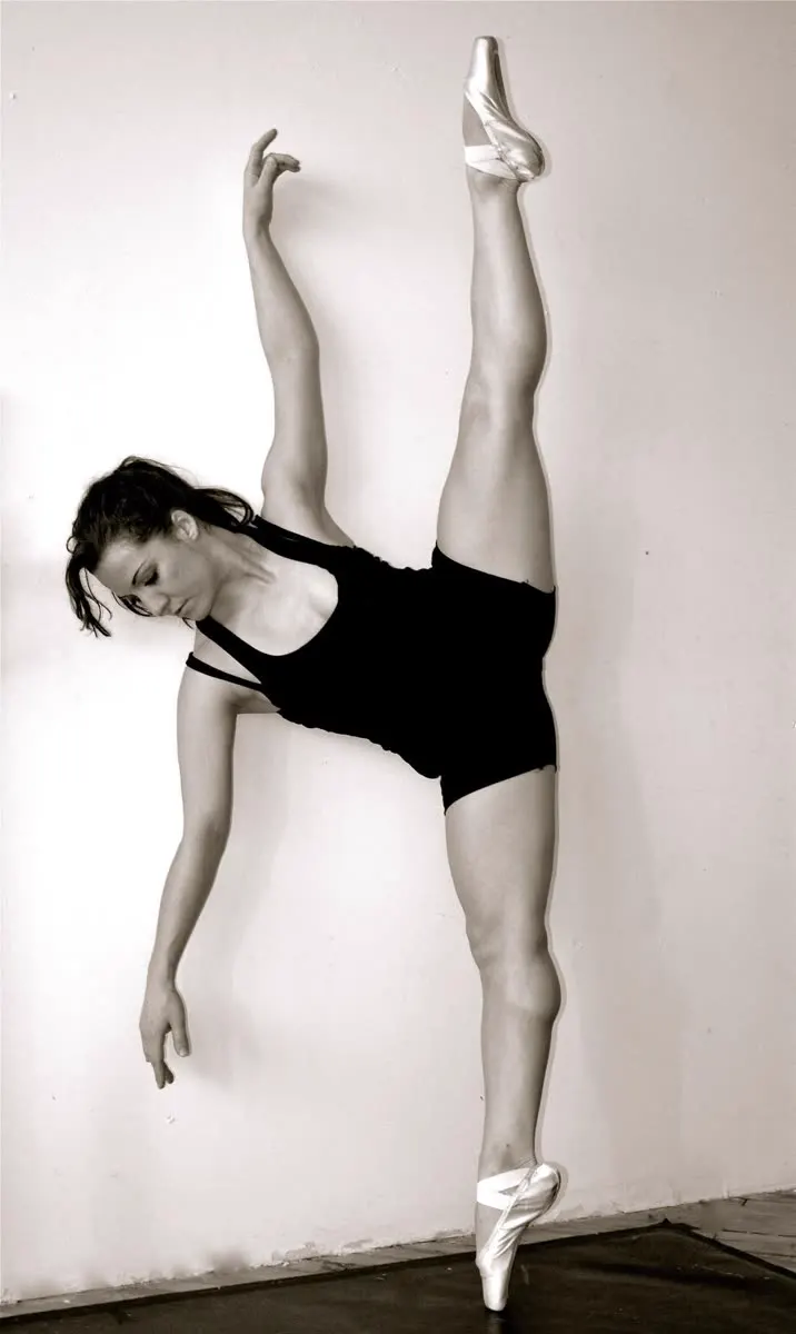 A dancer on pointe, one leg raised against a wall, in black and white